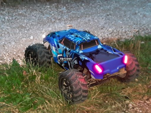 1-10 RC Monstertruck mit Lichtband Blauer RC-Monstertruck mit großen Reifen, steht im Gras auf einem Schotterweg.